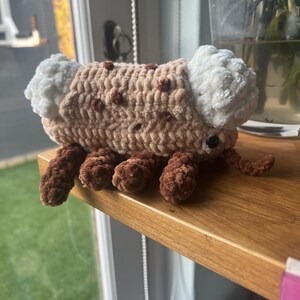 May include: A crocheted plush toy shaped like a cookie with cream filling, featuring a tan body with brown spots, white textured ends, dark brown legs, and small black eyes. The soft yarn construction creates a rounded, segmented appearance.