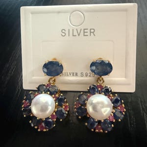 May include: A pair of gold-tone drop earrings featuring a central white pearl surrounded by a cluster of oval-cut blue sapphires and small round red rubies. Each earring has an oval blue sapphire stud top and is displayed on a white card labelled SILVER.