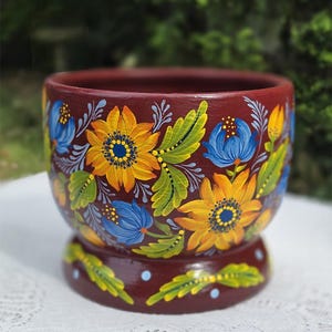 May include: A round, deep red ceramic planter featuring hand-painted yellow sunflowers, blue flowers, and green leaves. The base of the planter is also decorated with small green leaves and light blue dots, creating a cohesive floral design.