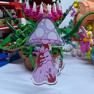 Könnte beinhalten: Ein durchsichtiger Acryl-Schlüsselanhänger mit einer hellvioletten Pilzillustration, dunkelroten Tropfenakzenten und einem kleinen Messermotiv. Der Pilzhut hat ein weißes Punktmuster und der Stiel zeigt ein Spiralmuster an der Basis.