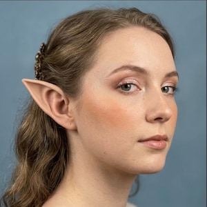 May include: A pair of prosthetic elf ears made of a smooth, skin-toned material. The ears feature a pointed, elongated shape that fits over the natural ear, blending with the skin to create a fantasy-inspired appearance for costume purposes.