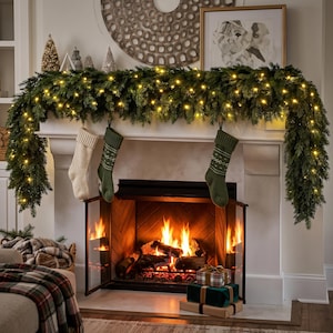 May include: A lush green artificial pine garland illuminated with warm white LED string lights, draped over a white fireplace mantel. Two stockings, one cream and one olive green with a white geometric pattern, hang from the garland.