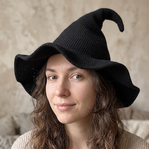 May include: A solid black knitted hat with a tall, pointed crown and a wide, wavy brim. The hat features a textured, ribbed stitch pattern throughout, with a distinct curve at the very tip of the pointed top, creating a classic conical silhouette.