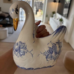 Może przedstawiać: Biała ceramiczna figurka łabędzia z ręcznie malowanymi niebieskimi wzorami kwiatowymi na korpusie i skrzydłach. Łabędź ma długą, wygiętą szyję, złoty dziób i szczegółowe niebieskie oczy. Powierzchnia ceramiki jest gładka i błyszcząca.