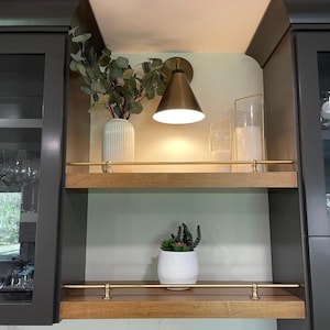 May include: Two horizontal wooden shelves with brass railings mounted between dark gray cabinets. The top shelf holds a ribbed white vase with eucalyptus, a brass cone wall sconce, and two glass candle holders. The bottom shelf holds a white pot with succulents.