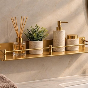 Puede incluir: Una estantería de pared rectangular de metal dorado con una barra protectora frontal. La estantería sostiene un difusor de varillas de cristal, una pequeña planta verde en una maceta beige moteada, un dispensador de jabón y un tarro a juego.