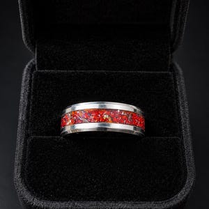 Puede incluir: Un anillo de metal plateado con una incrustación central ancha de material rojo y naranja triturado con una textura brillante y centelleante. La banda tiene bordes plateados pulidos y se presenta dentro de un joyero forrado de terciopelo negro.