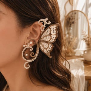 Peut inclure: Une boucle d'oreille en forme d'aile de papillon crochetée avec du fil crème et marron. La pièce présente des accents complexes en forme de spirale et de petites perles blanches sphériques intégrées à la structure, couvrant le lobe et le cartilage.