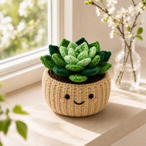 Puede incluir: Una planta suculenta de ganchillo con hojas verdes en capas en una maceta beige, redonda y acanalada. La maceta presenta una cara bordada con dos ojos negros circulares, una pequeña boca negra curvada y dos mejillas rosadas pequeñas.