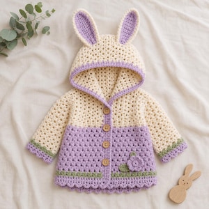 May include: A cream and lavender crochet hooded cardigan featuring long sleeves, a hood with two tall bunny ears, four brown wooden buttons down the center, and a small crocheted flower detail on the lower right side with a green trim.