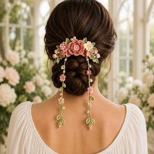Puede incluir: Un accesorio para el cabello con un grupo de flores de ganchillo en tonos rosa, crema y rosa viejo, acentuado con hojas verdes de ganchillo y pequeñas perlas blancas. Dos largas hebras colgantes de flores y hojas de ganchillo cuelgan.