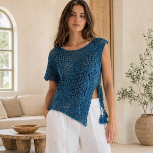 May include: A deep blue crochet top featuring an intricate openwork pattern with fan-like motifs, a wide neckline, and an asymmetrical hemline. The side is secured with matching blue cord ties that end in decorative tassels with small white beads.