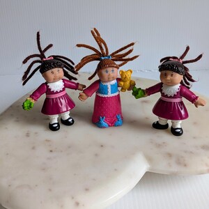 May include: Three small plastic dolls with yarn hair. Two dolls wear magenta dresses with white collars and black shoes. The center doll wears a blue vest over a pink dress with blue rabbit accents and holds a small yellow bear figure.