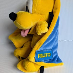 May include: A yellow plush dog toy with a black nose and a pink tongue, wrapped in a blue blanket with a yellow border. A rectangular blue patch on the blanket features the name PLUTO embroidered in yellow thread, with a black tail extending below.
