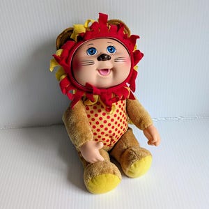 Op de afbeelding: Een pluche pop met een vinyl gezicht met blauwe ogen, een kleine neus en een lachende mond. De pop draagt een rood-gele leeuwenkap met manen en een beige lichaam met rode stippen, met gele stof op de voeten.