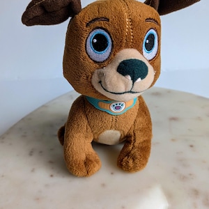 May include: A brown plush dog toy with large blue eyes, a dark brown nose, and floppy ears. The dog wears a light blue collar featuring a tan band-aid design and a central circular emblem with a purple paw print graphic on a white background.