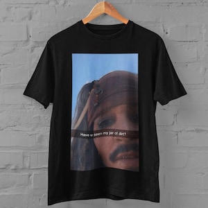 Op de afbeelding: Een zwart t-shirt met korte mouwen en een rechthoekige grafische print van een persoon met een bruine hoofddoek tegen een blauwe lucht. De tekst "Have u seen my jar of dirt?" staat in het wit gedrukt in het midden van de afbeelding.