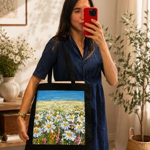 Könnte beinhalten: Eine schwarze Tragetasche mit einem rechteckigen, strukturierten Ölgemälde-Druck eines lebendigen Feldes voller weißer Gänseblümchen unter einem hellblauen Himmel. Die Tasche hat zwei schwarze Stoffträger und eine schlichte, rechteckige Form.