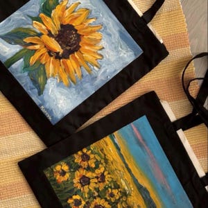 Könnte beinhalten: Zwei schwarze Tragetaschen mit aufgemalten Sonnenblumenmotiven. Die obere Tasche zeigt eine einzelne große Sonnenblume vor hellblauem Hintergrund. Die untere Tasche zeigt ein Sonnenblumenfeld unter einem blauen und rosafarbenen Himmel.