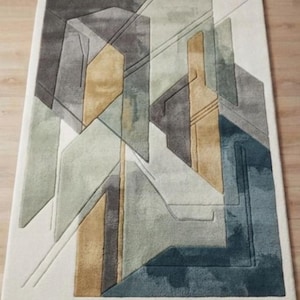Puede incluir: Una alfombra rectangular con un patrón geométrico de formas superpuestas en tonos gris carbón, verde salvia apagado, amarillo mostaza y azul cerceta, sobre un fondo blanco roto con detalles de textura tallada en la superficie.