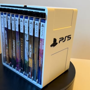 Puede incluir: Un estante de almacenamiento de plástico blanco para cajas de juegos, con el logotipo negro de PS5 en el lateral. El estante sostiene diez cajas de juegos verticales con lomos azules, mostrando claramente la marca PS5 en la parte superior.