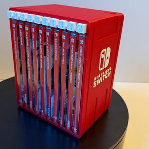 May include: A red plastic Nintendo Switch game storage case holding ten game cartridges. The case features the white Nintendo Switch logo on the side and includes ten small white pull tabs at the top for each individual game slot, allowing for easy access to the collection.