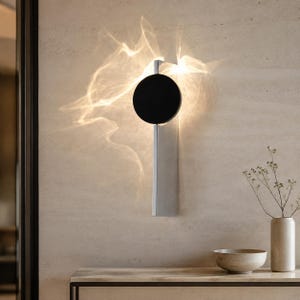 Op de afbeelding: Een wandlamp met een verticale rechthoekige zilveren basis en een cirkelvormige zwarte schijf in het midden aan de bovenkant. Het licht straalt een heldere, vloeiende, golfachtige gloed uit die de omgeving verlicht met een zachte warmte.