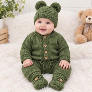 Peut inclure: Une tenue de bébé tricotée vert sauge composée d'une grenouillère à manches longues boutonnée avec une taille à cordon et des chaussons assortis, accompagnée d'un bonnet texturé orné de deux pompons sur le dessus, en laine douce.