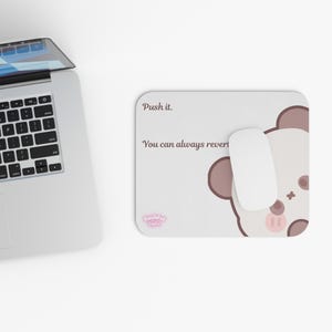 May include: A rectangular mouse pad with a light beige surface featuring a brown cartoon bear face, a white computer mouse, and the text 'Push it. You can always revert'. A silver laptop is positioned to the left of the mouse pad.