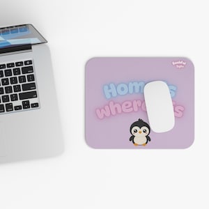 以下が含まれることがあります： ライトパープルの背景に漫画のペンギンと青とピンクのバブル文字で「Home is where I is」と書かれた長方形のマウスパッド。白いコンピュータマウスがパッドの上に置かれており、「Bashful Byte」のロゴも表示されています。
