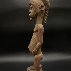 Pode incluir: Uma escultura de madeira esculpida à mão de uma figura em pé com um abdômen proeminente, cabelo texturizado e padrões geométricos gravados nas bochechas e no estômago. A figura é retratada de perfil, mostrando uma barriga arredondada e traços.