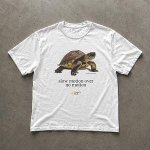 May include: A white short-sleeve t-shirt featuring a realistic brown and green turtle graphic. Below the turtle, the text reads "slow motion over no motion" in black, followed by a small yellow logo and the text "Launch Club".