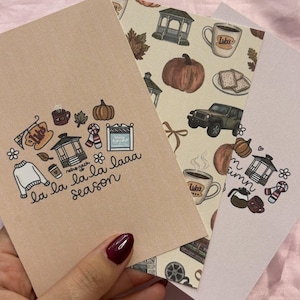 Peut inclure: Trois cartes en papier rectangulaires avec des illustrations sur le thème de l'automne. La carte avant est beige avec des icônes de pull, tasse de café, kiosque et citrouille, avec le texte "la la la la laa season". D'autres cartes montrent des tasses.