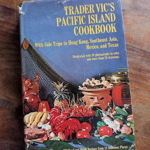 May include: A hardcover book titled Trader Vic's Pacific Island Cookbook with a blue cover featuring a colorful illustration of a lobster, pineapple, tomatoes, and a ceramic tiki-style bowl. The text includes side trips to Hong Kong and Texas.