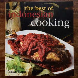 Pode incluir: Um livro de culinária intitulado The Best of Indonesian Cooking de Yasa Boga. A capa apresenta uma fotografia de um peixe frito inteiro coberto com um molho espesso de pimenta sambal vermelho vibrante, servido num prato branco com coentros.
