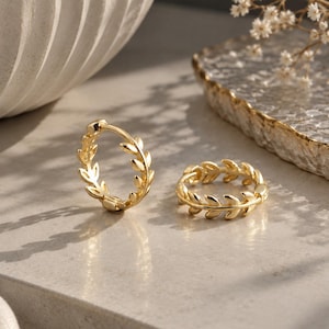 May include: A pair of gold hoop earrings featuring a continuous leaf or vine pattern along the outer edge. One earring stands upright while the other lies flat, both showcasing a polished metallic finish and a hinged closure mechanism.