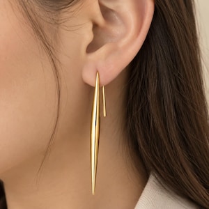 May include: A long, slender, gold-toned earring with a smooth, polished finish. The earring features a pointed, needle-like shape that hangs vertically from a simple, thin wire hook that passes through the earlobe, creating a sleek look.