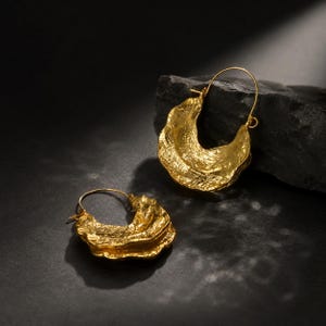 Peut inclure: Une paire de créoles dorées présentant une surface texturée, irrégulière et organique. Les boucles d'oreilles ont un design en forme de croissant avec une finition métallique polie qui capte la lumière, créant un aspect martelé distinct.
