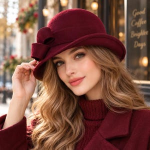 Puede incluir: Un sombrero cloche de fieltro color burdeos intenso con un ala ancha y estructurada y un detalle de lazo decorativo prominente en el lateral. El sombrero está confeccionado en un material liso de color sólido y se lleva con un suéter de cuello alto a juego.