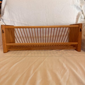 May include: A wall-mounted wooden rack featuring a horizontal base and a series of vertical slats angled outward to create a storage compartment. The unit is constructed from light-toned wood and includes two keyhole slots on the side panels for mounting.