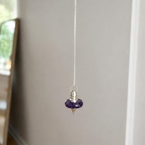 May include: A purple faceted gemstone pendulum suspended from a thin silver-colored metal chain. The pendulum features a polished, dark purple stone bead shaped like a flattened sphere, capped with a silver-colored metal top and a pointed tip.