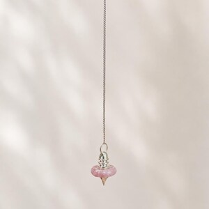 Könnte beinhalten: Eine silberfarbene Metallkette hält ein spitzes Pendel aus einem rosafarbenen Edelstein. Der Anhänger besteht aus einer glatten, kreisförmigen rosa Steinperle, die von einer polierten silberfarbenen Metallkappe gekrönt wird, die in einer Spitze endet.