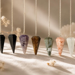 May include: Seven faceted crystal pendulums hanging from silver-toned chains. The stones include black, purple amethyst, brown tiger's eye, green moss agate, peach sunstone, and pale white quartz, each shaped into a pointed geometric form.