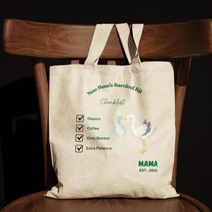 Könnte beinhalten: Eine beige Stofftasche aus Segeltuch mit dem grünen Text New Mom's Survival Kit Checklist. Darunter befinden sich vier abgehakte Kästchen mit den Bezeichnungen Diapers, Coffee, Cozy Blanket und Extra Patience. Eine Storchenillustration und MAMA EST. 2026.
