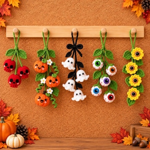 May include: Five hanging crochet ornaments featuring red skulls, orange pumpkins with white flowers, white ghosts on a black vine, multi-colored eyeballs with green leaves, and yellow sunflowers with green leaves, all suspended from a wooden wall rack.