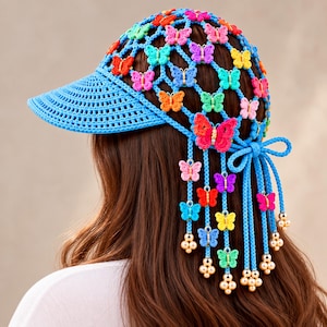 May include: A blue crocheted cap featuring a lattice pattern adorned with small, colorful butterfly embellishments. The cap includes a solid blue brim and long, dangling blue cords decorated with additional butterflies and small cream-colored beads.