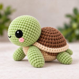 May include: A small, handmade crochet turtle toy featuring a light green head and limbs, a dark brown shell, and a beige underside. The turtle has large, glossy black safety eyes, a small embroidered black mouth, and round pink stitched cheeks.
