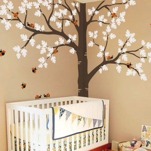 Könnte beinhalten: Ein weißes Babybett mit braunem Fuß und einer weißen und blauen Bettdecke. Das Bett steht in einem Kinderzimmer mit einer braunen Wand und einem großen weißen Baum-Aufkleber mit braunen Ästen und roten Marienkäfern. Der Aufkleber befindet sich an der Wand hinter dem Bett. Die Wand hat auch eine weiße Alphabettafel mit bunten Buchstaben. Der Text auf der Tafel lautet "ABCDEFGHIJKLMNOPQRSTUVWXYZ".