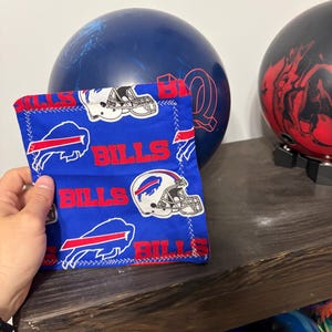 May include: A square fabric pouch featuring a repeating pattern of the Buffalo Bills logo, including the blue bison silhouette and white football helmet with red and blue accents, set against a bright blue background with red text reading BILLS.
