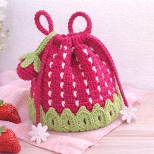 May include: A crochet drawstring bag shaped like a strawberry, featuring a deep red body with vertical white stitched lines, a light green scalloped base, two top handles, and a small strawberry charm attached to a green cord with white flower ends.